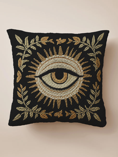A black woven throw pillow with intricate gold and ivory embroidery featuring an Evil Eye motif, surrounded by botanical flourishes and sunburst details.